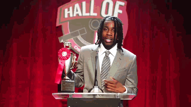 a man in a suit and tie is giving a speech in front of a sign that says hall of