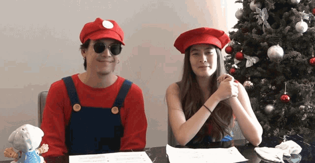 a man dressed as mario and a woman dressed as chef are sitting at a table in front of a christmas tree