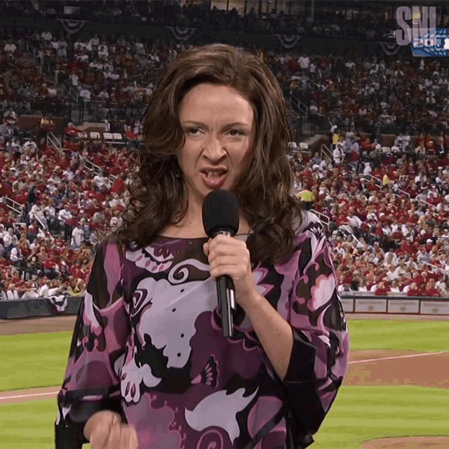 a woman in a purple shirt is holding a microphone in front of a crowd that is watching a baseball game