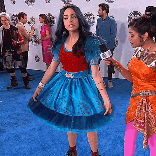 a woman in a blue dress is being interviewed on a blue carpet with neon lights written on the wall behind her