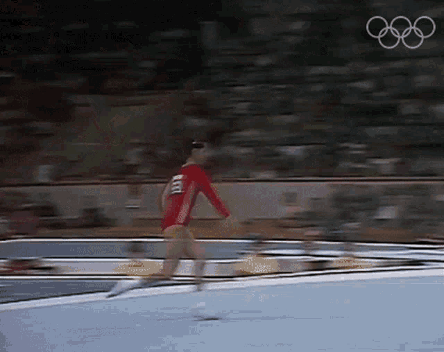 a woman in a red leotard is doing a trick on a trampoline with the olympics logo in the background