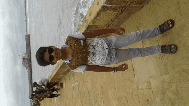 a boy wearing sunglasses stands on a brick wall near the ocean