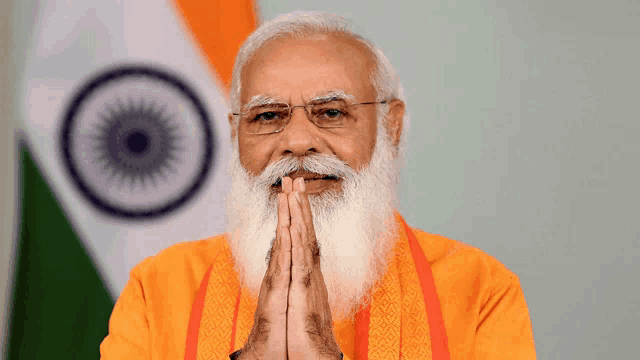 a man with a beard and glasses holds his hands together in front of the indian flag