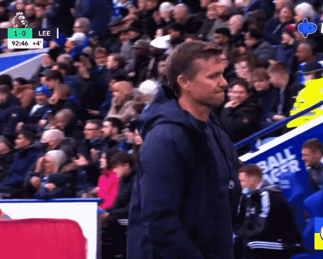 a man in a blue jacket stands in front of a crowd at a soccer game with a score of 1-0