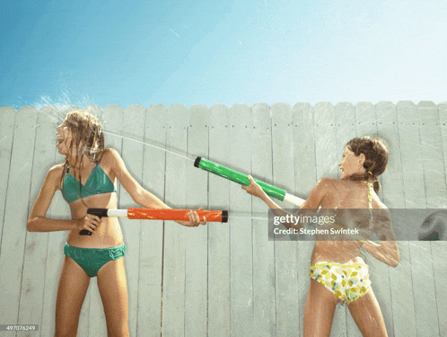 two girls in bikinis playing with water guns on a fence