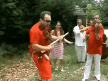 a man in a red shirt is playing a guitar in front of a group of people in white pants .