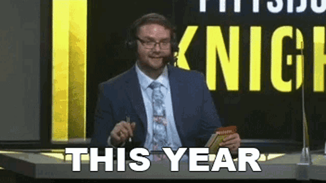 a man in a suit and tie is sitting in front of a sign that says " this year "