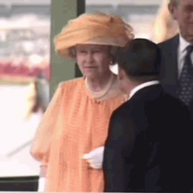 the queen is wearing an orange dress and a yellow hat while talking to a man in a suit .