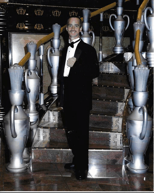 a man in a tuxedo stands on a set of stairs with silver vases