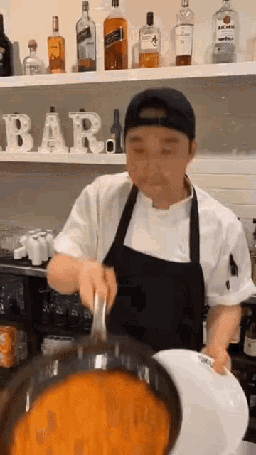 a man in an apron is cooking in a kitchen with bottles of alcohol on the shelves behind him .