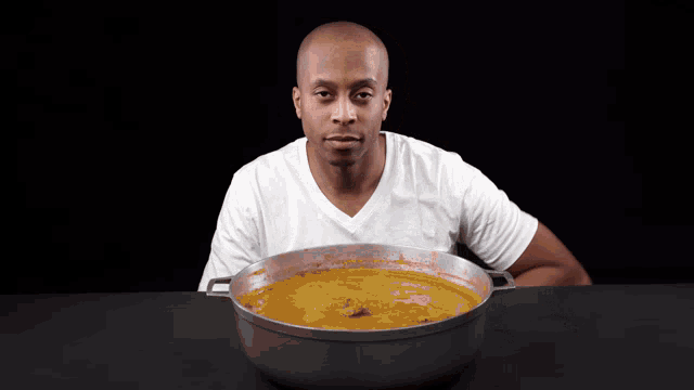 a man in a white shirt is sitting at a table with a pot of soup in front of him