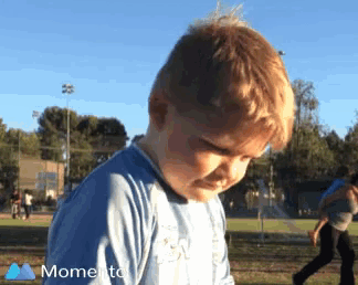 a young boy in a blue shirt is standing in a park with a moments logo on the bottom right