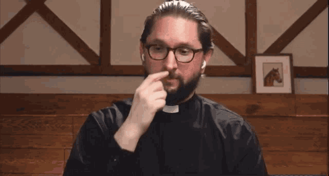 a man wearing glasses and a priest collar holds his hand to his nose