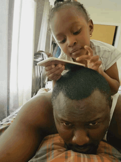 a little girl sits on a man 's head looking at his phone