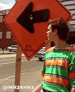 a man in a green and orange striped shirt stands in front of an orange sign with an arrow pointing right