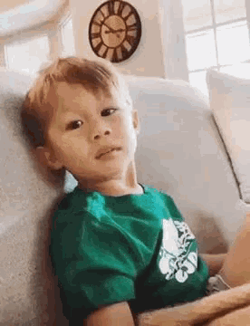 a young boy in a green shirt is sitting on a couch with a clock on the wall behind him .