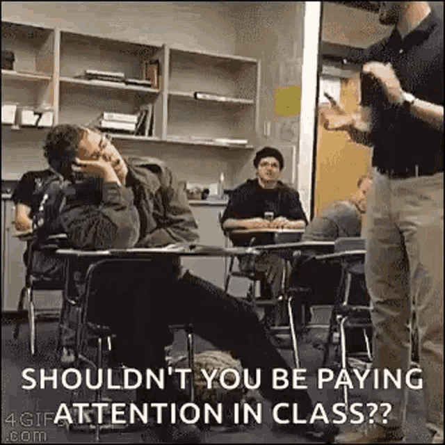 a man is sitting at a desk in a classroom while a teacher talks to a group of students .