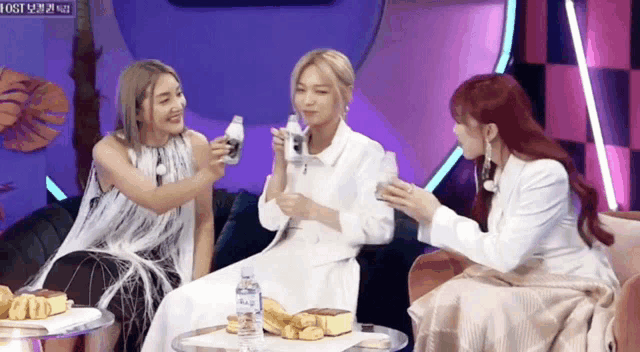 three women are sitting on a couch holding bottles and drinking water .