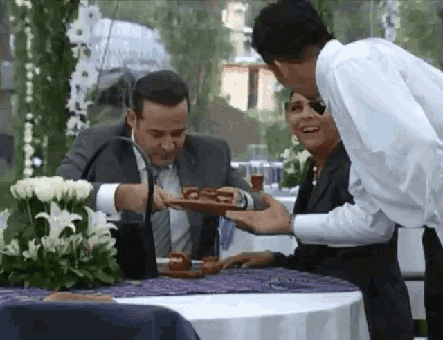 a man and woman are sitting at a table with flowers and a man is holding a plate of food