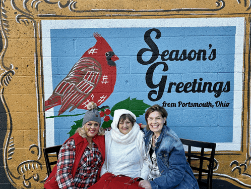 three women are posing in front of a mural that says season 's greetings from portsmouth ohio