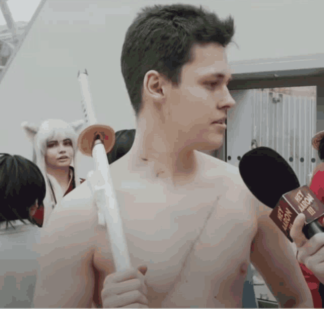 a shirtless man holding a sword is being interviewed by a microphone that says next arrow