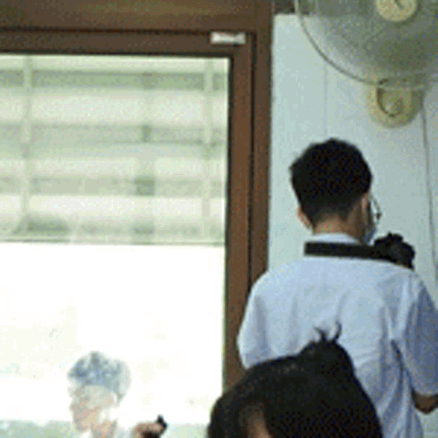 a man in a white shirt is standing in front of a window with a fan on the wall behind him