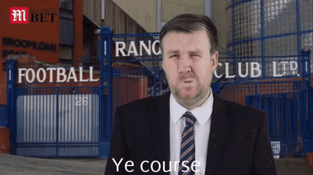 a man in a suit and tie stands in front of a fence that says ' rangers club ltd ' on it