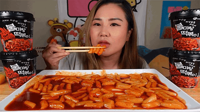 a woman is eating rice cakes with chopsticks in front of a stack of cups that say ' korean food ' on them