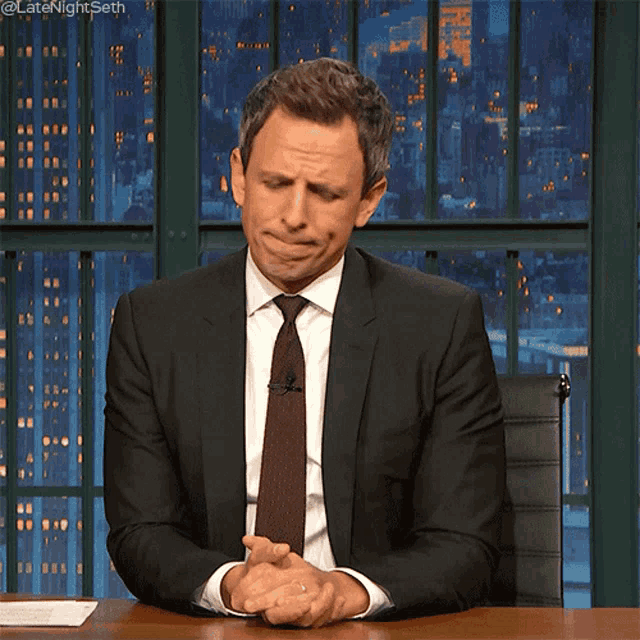 a man in a suit and tie is sitting at a desk with his hands folded in front of him