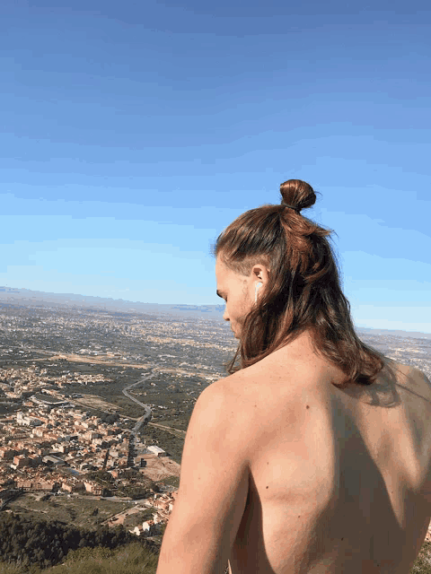 a shirtless man with long hair in a bun looks at a city