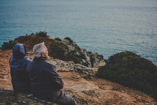 An elderly couple sit on a rocky beach looking out to the sea.