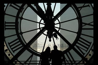 Silhouettes of two people in front of the inside of a large, clear clock overlooking a city.