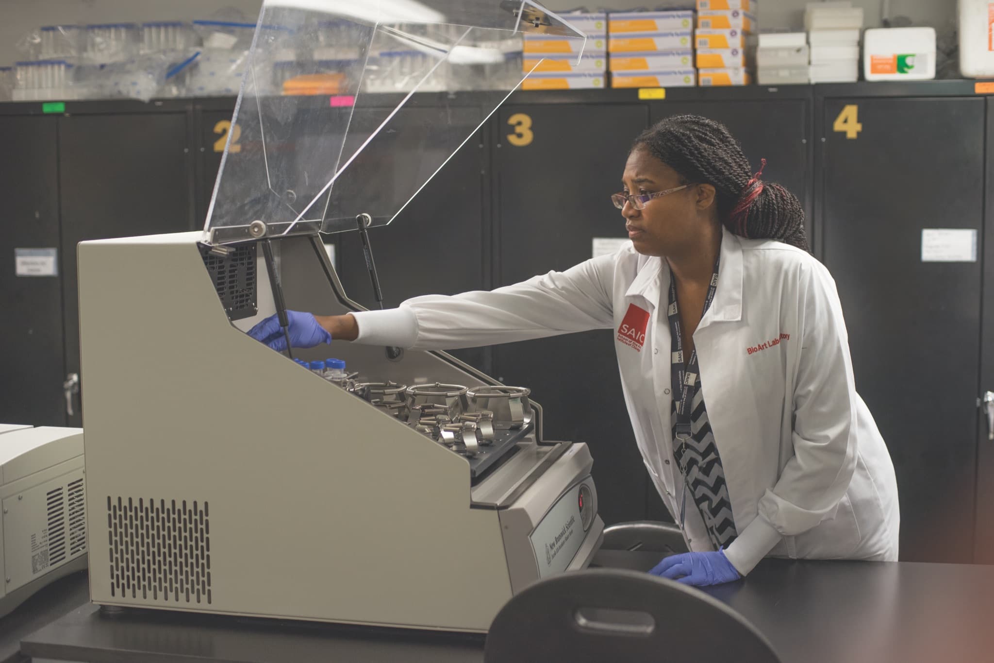 A young woman wearing a white lab coat and blue rubber gloves leans on a table and into a piece of lab equipment.