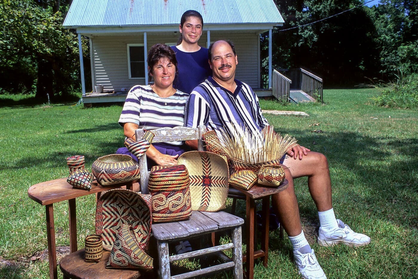 Three Chitimacha Triberibal members, one standing and two seated with some of their baskets on wooden furniture in front of a small house.