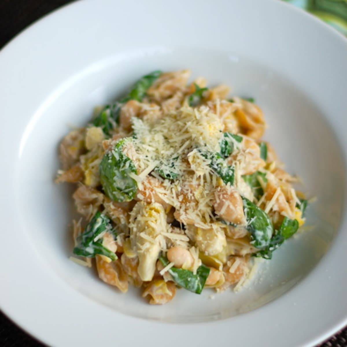 Spinach artichoke pasta with cheese topping in a white bowl.