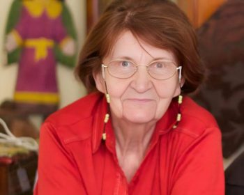 Portrait of Professor Alicja Kuczyńska, lady in a red shirt with art pieces in the background, blurred.