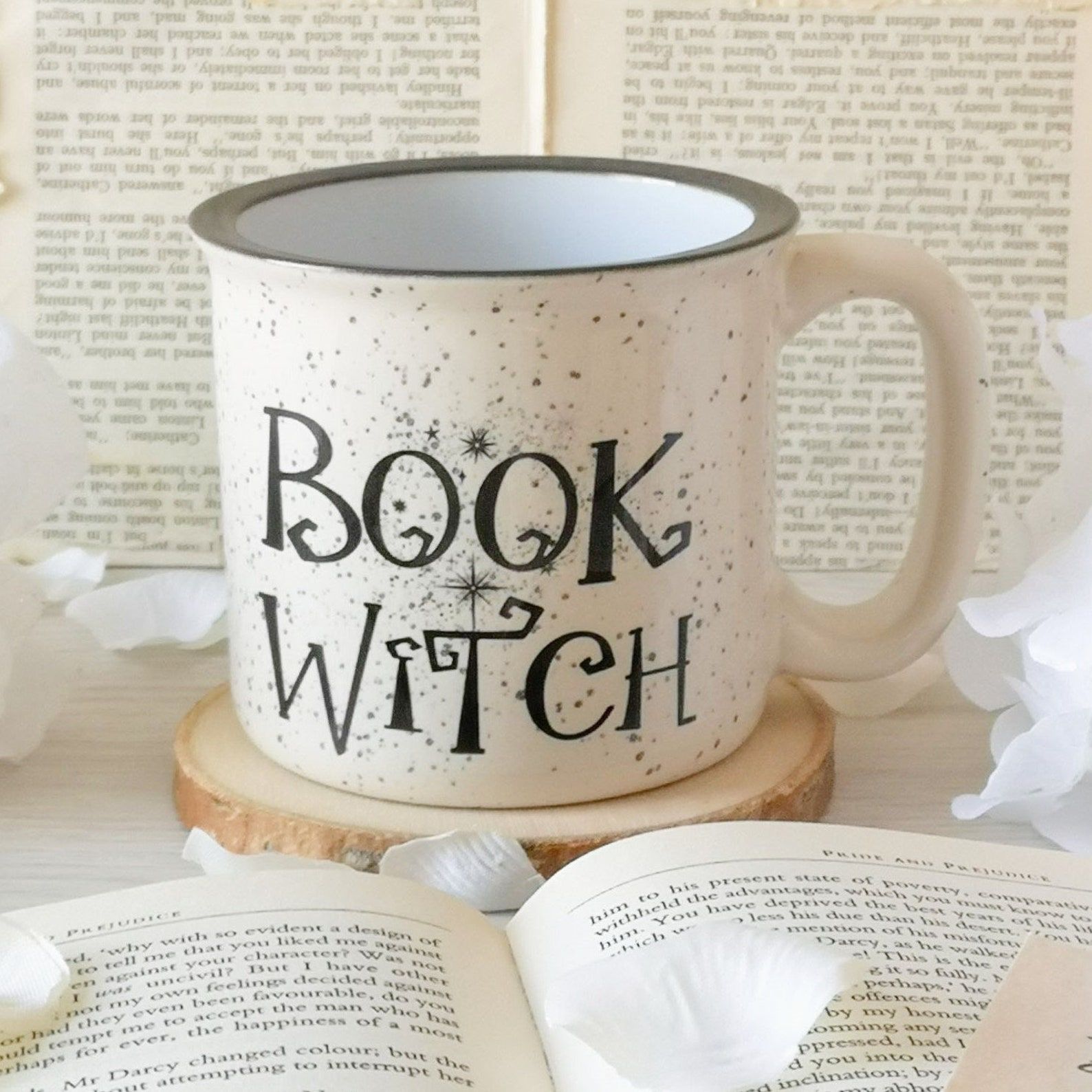 Image of a white flecked mug on a coaster in front of an open book. The mug reads "book witch." 