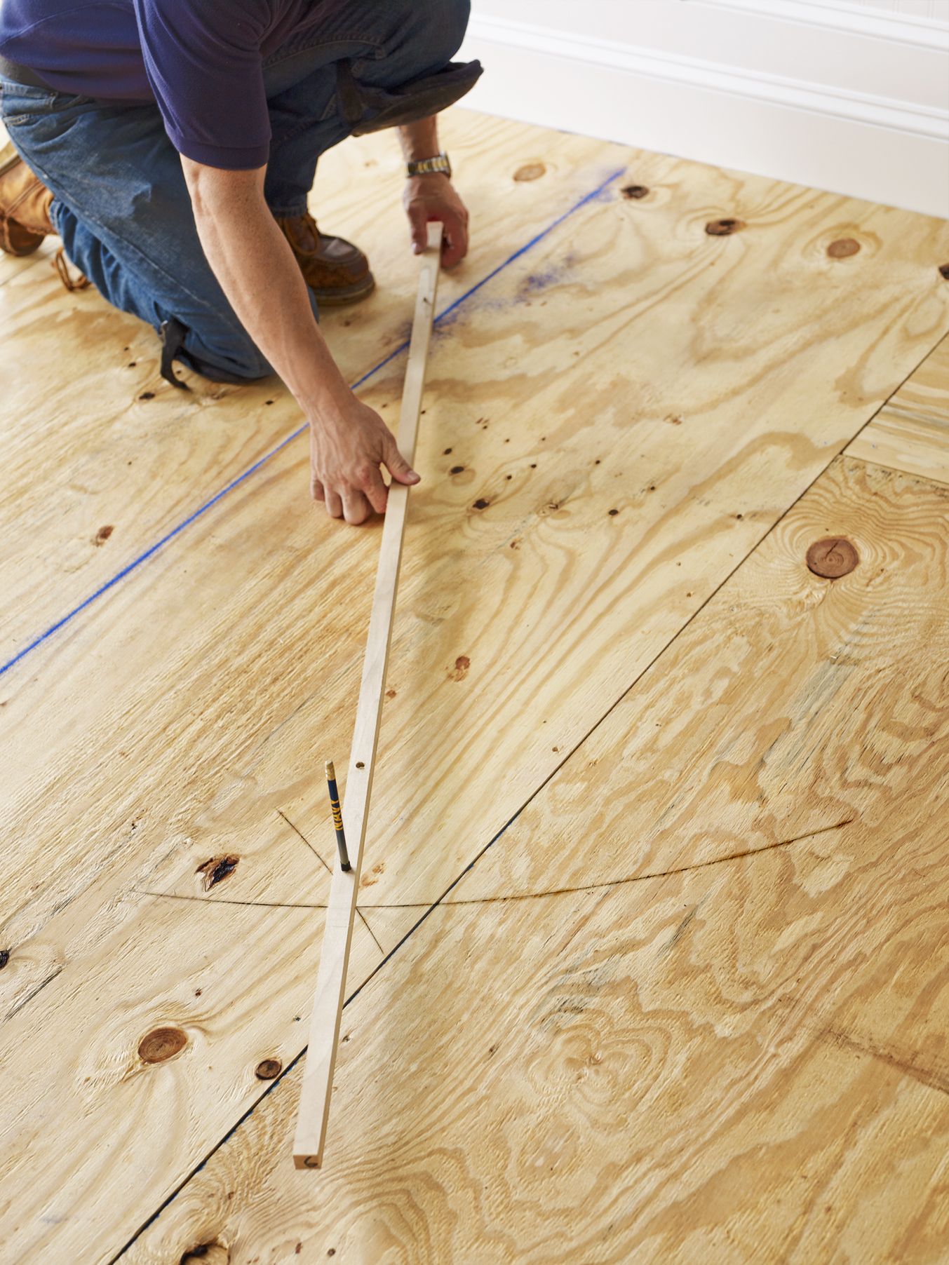 A person uses a trammel to draw a semicircle across the room's centerline to mark the ground for installing the herringbone floor.