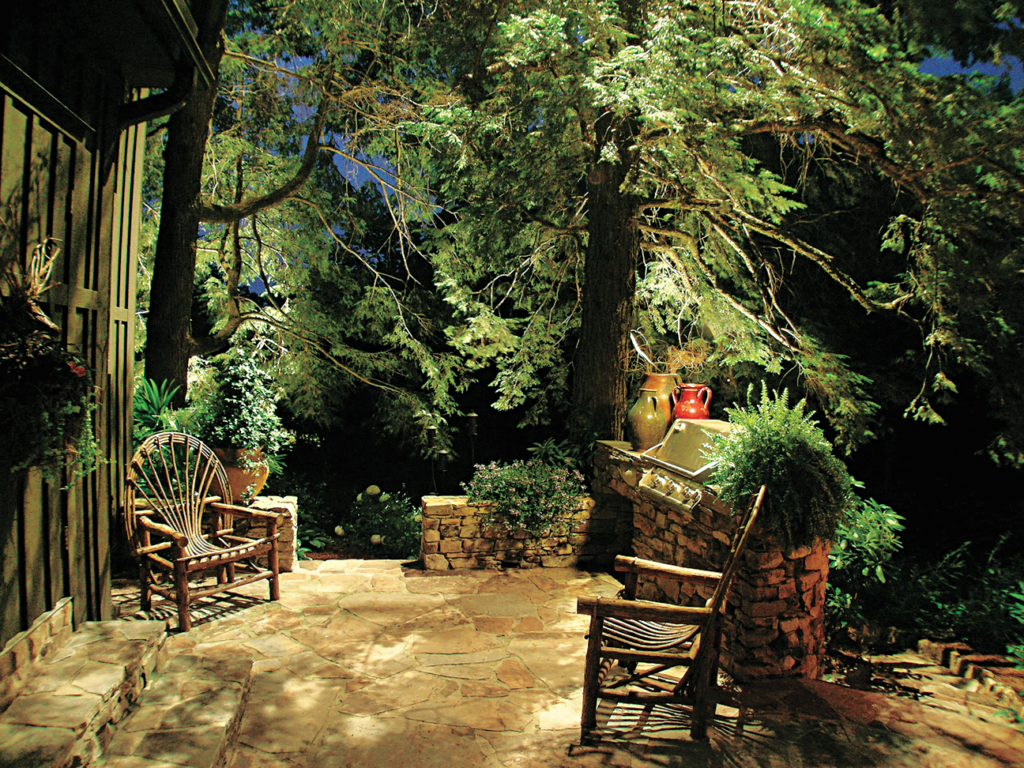 A stone patio lit by landscape lighting, it has two chairs and a grill.