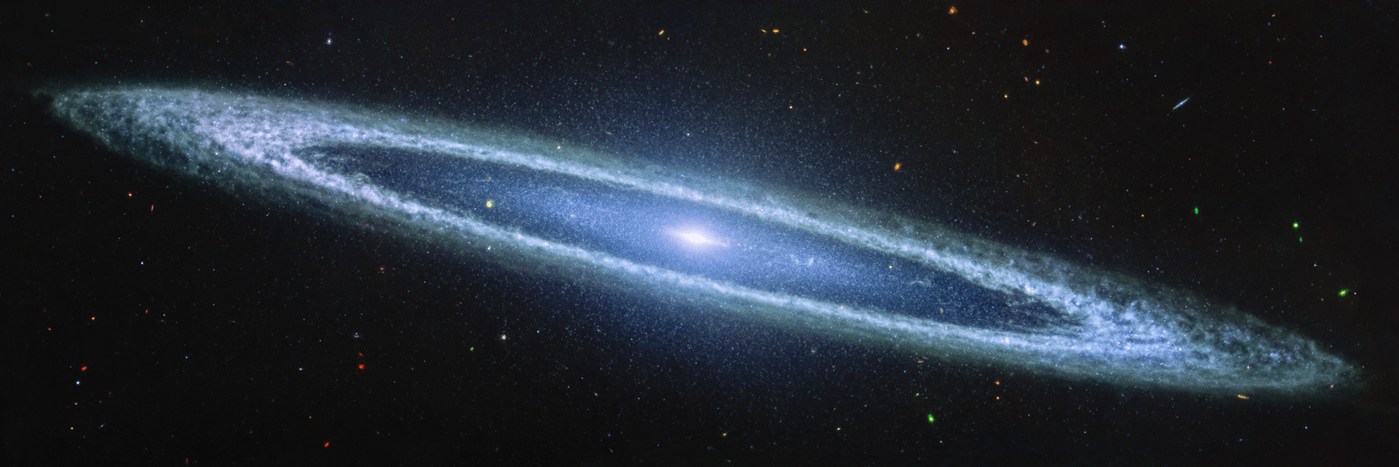 Image of a galaxy on the black background of space. The galaxy is a very oblong, blue disk that extends from left to right at an angle (from about 10 o’clock to 5 o’clock). The galaxy has a small bright core at the center. There is an inner disk that is clearer, with speckles of stars scattered throughout. The outer disk of the galaxy is whiteish-blue, and clumpy, like clouds in the sky. There are different colored dots, distant galaxies, speckled among the black background of space surrounding the galaxy.