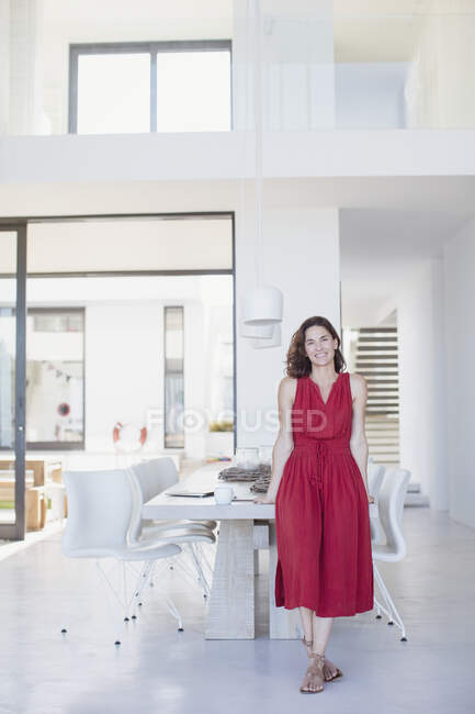 Porträt lächelnde brünette Frau in rotem Kleid lehnt am Esstisch — Stockfoto