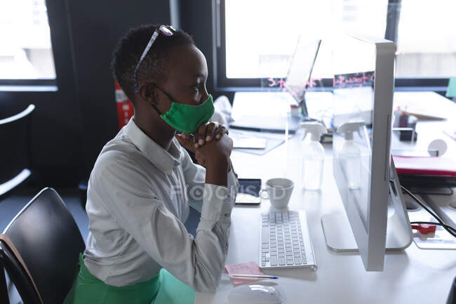 Afroamerikanerin mit Gesichtsmaske blickt im modernen Büro auf ihren Computerbildschirm. Soziale Distanzierung von Quarantäne während der Coronavirus-Pandemie — Stockfoto