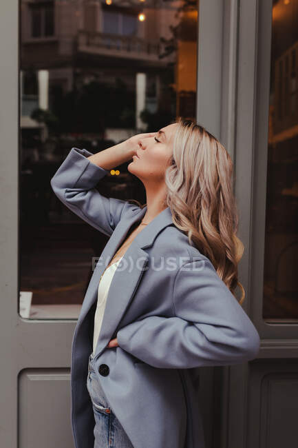 Seitenansicht einer Frau mit langen blonden Haaren und in stylischem Outfit, die mit geschlossenen Augen in der Nähe eines Gebäudes in der Stadt steht — Stockfoto