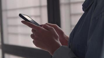 Asian business woman holding typing mobile phone and scrolls through social media feed in smartphone standing beside window at home office. video