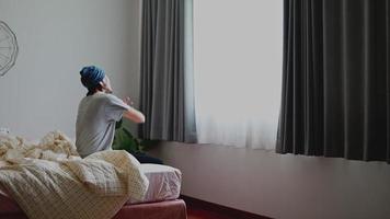 Asian man waking up and stretches while sitting on the bed in bedroom morning sun shines through the window at home. video