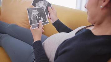 Happy Asian pregnant woman watching ultrasound image on sofa. video