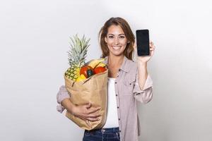 Young woman holding paper bag full of groceries and showing her mobile phone, Order online concept. shopping app photo