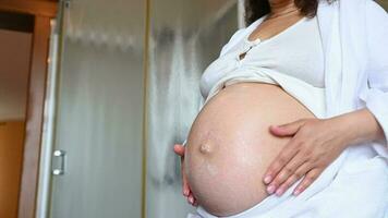 Midsection of a gravid expectant woman massaging her pregnant belly, rubbing stretch marks cosmetics in the home bathroom. Home spa procedures, Skin and body care in pregnancy time. Hygiene concept video