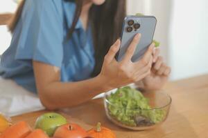 A young woman with a beautiful face in a blue shirt with long hair eating fruit sitting inside the kitchen at home with a laptop and notebook for relaxation, Concept Vacation. photo