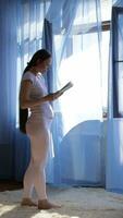 a pregnant woman in white pants and a white shirt is standing in front of a window reading a book video